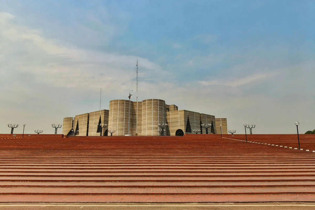 The National Parliament House in Dhaka under a clear blue sky, above its wide red brick steps