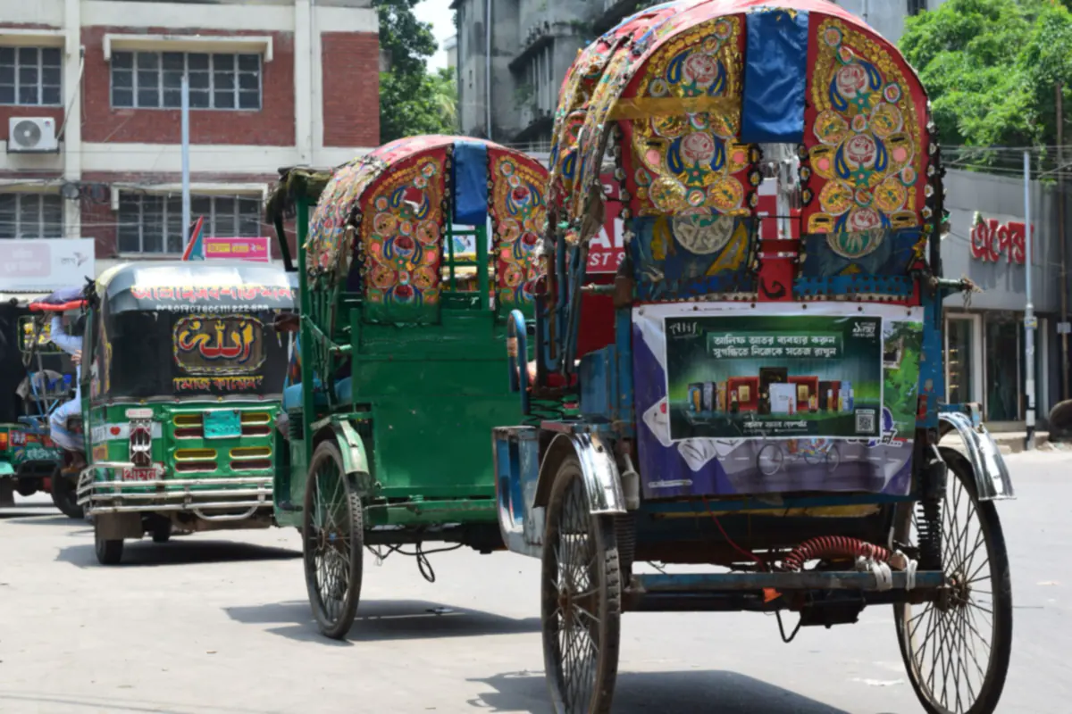 Two cycle rickshaws with hoods painted in red, gold and blue flowers, next to a green auto rickshaw on a Dhaka street
