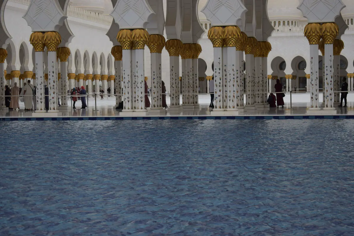 White columns with floral inlay and gold capitals beside a blue pool in the courtyard of the Sheikh Zayed Grand Mosque