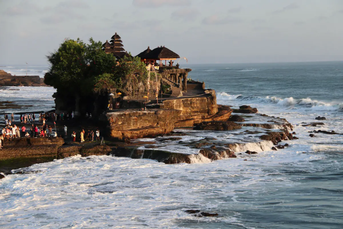 Tanah Lot temple with tiered shrine roofs on a rock outcrop, waves breaking around it and visitors on the shore