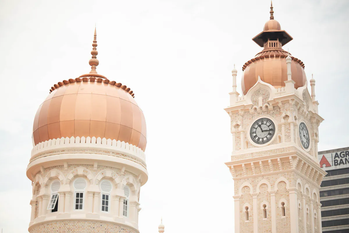 A copper dome and the clock tower of the Sultan Abdul Samad Building in Kuala Lumpur against a pale sky