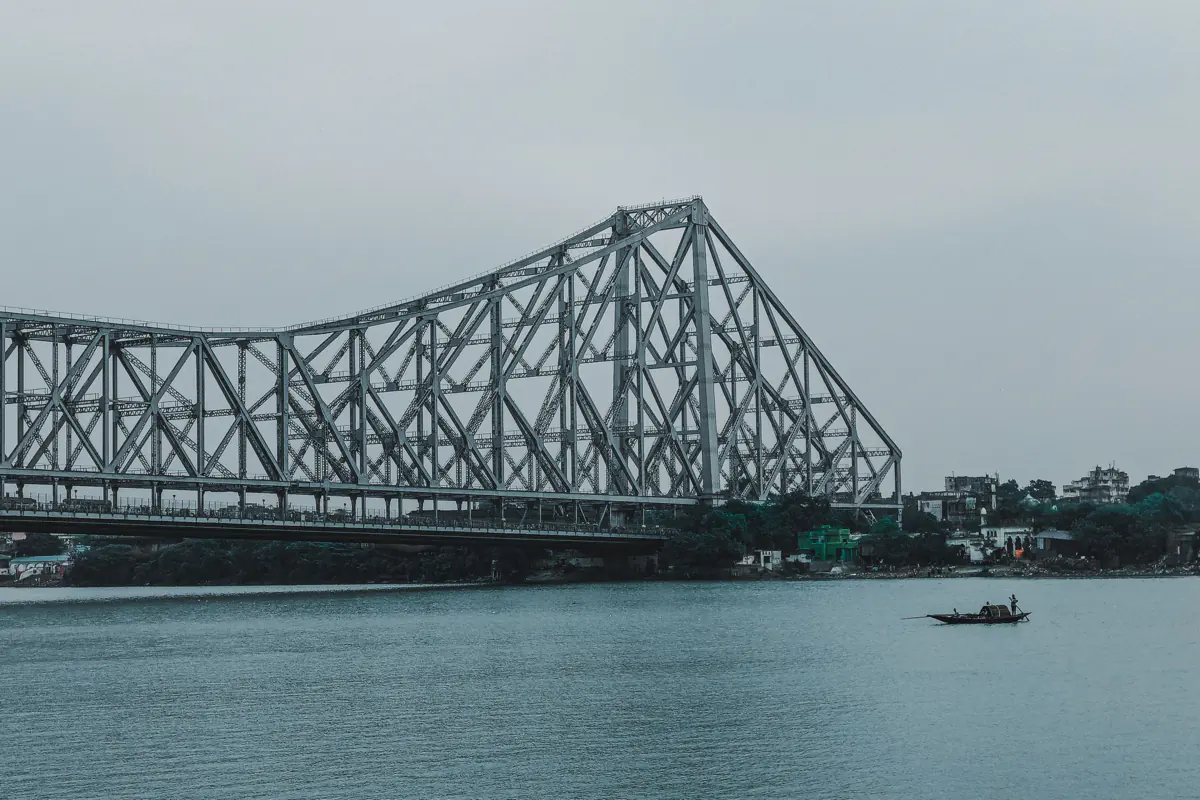 The steel lattice of Howrah Bridge over the Hooghly River in Kolkata, with a small wooden boat on the water below