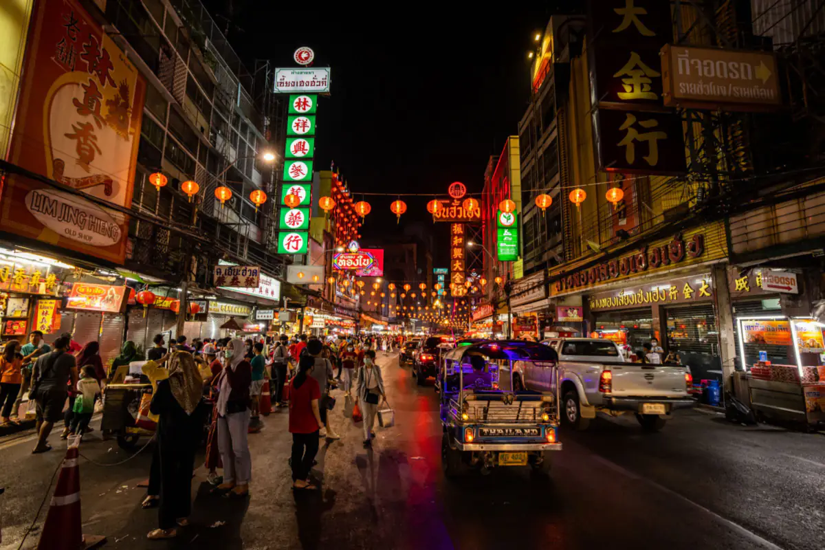 A street in Bangkok's Chinatown at night, with neon signs, red lanterns, people walking and a tuk tuk on the road