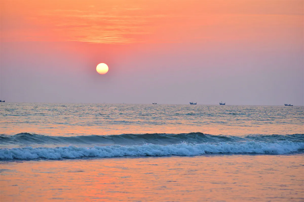 The sun setting over the Bay of Bengal, with waves breaking in the foreground and fishing boats on the horizon