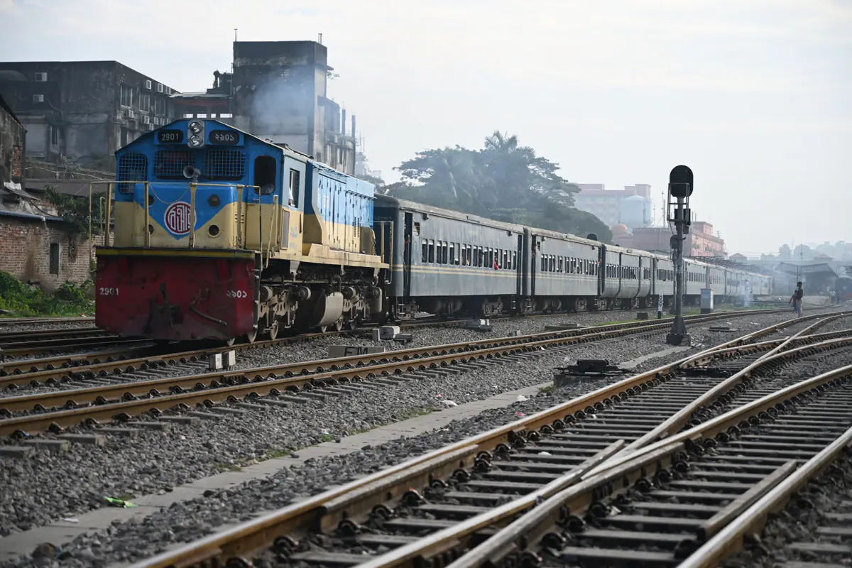 A blue and yellow diesel locomotive pulling passenger coaches past the tracks at Chattogram station