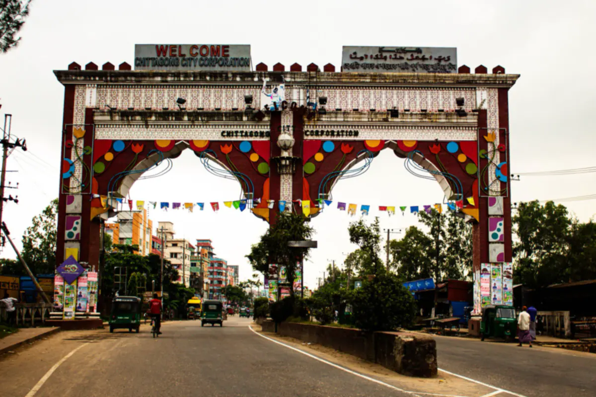 A colourful city entrance arch over a road in Chattogram, with auto rickshaws and a cyclist passing beneath