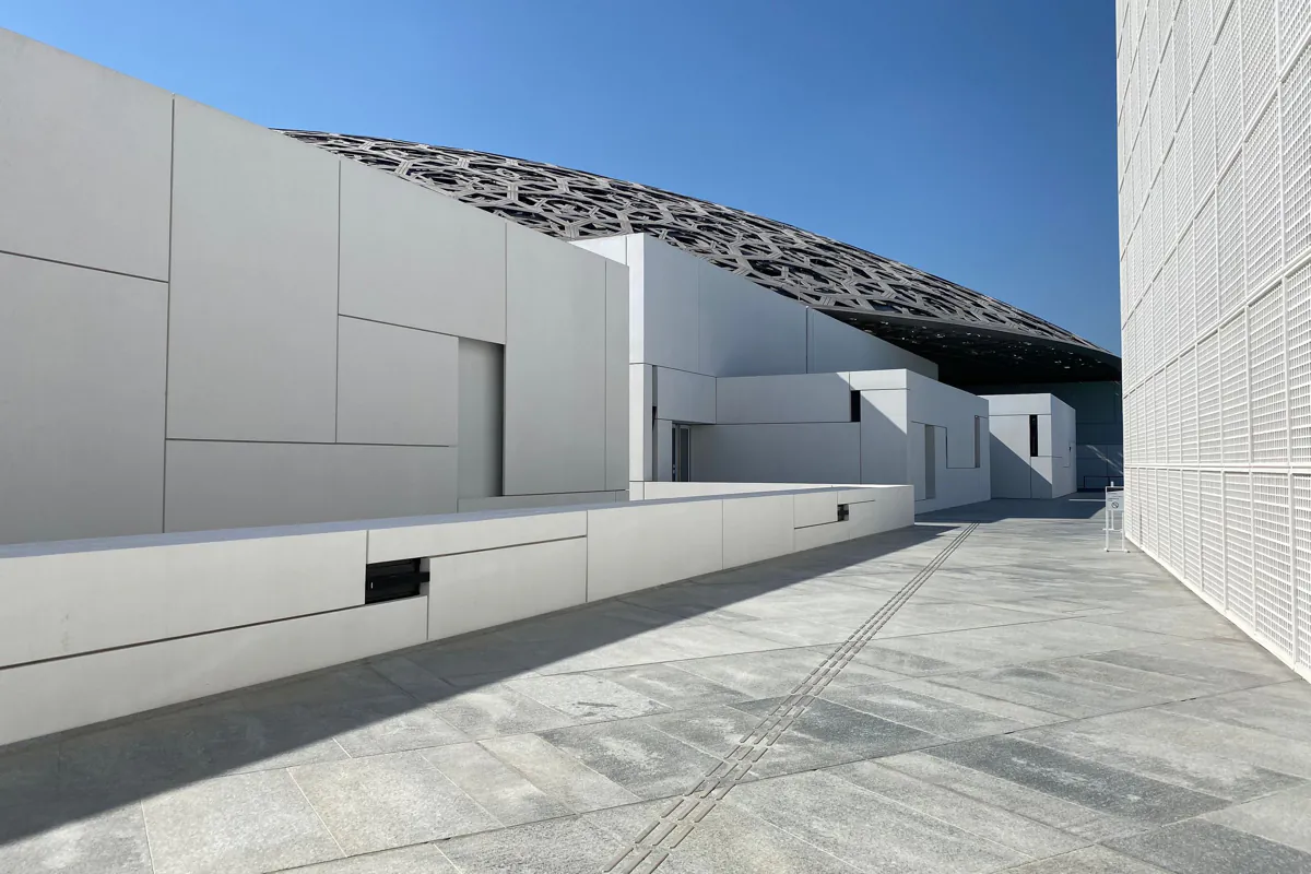 White gallery blocks and a paved terrace beneath the patterned metal dome of Louvre Abu Dhabi under a blue sky