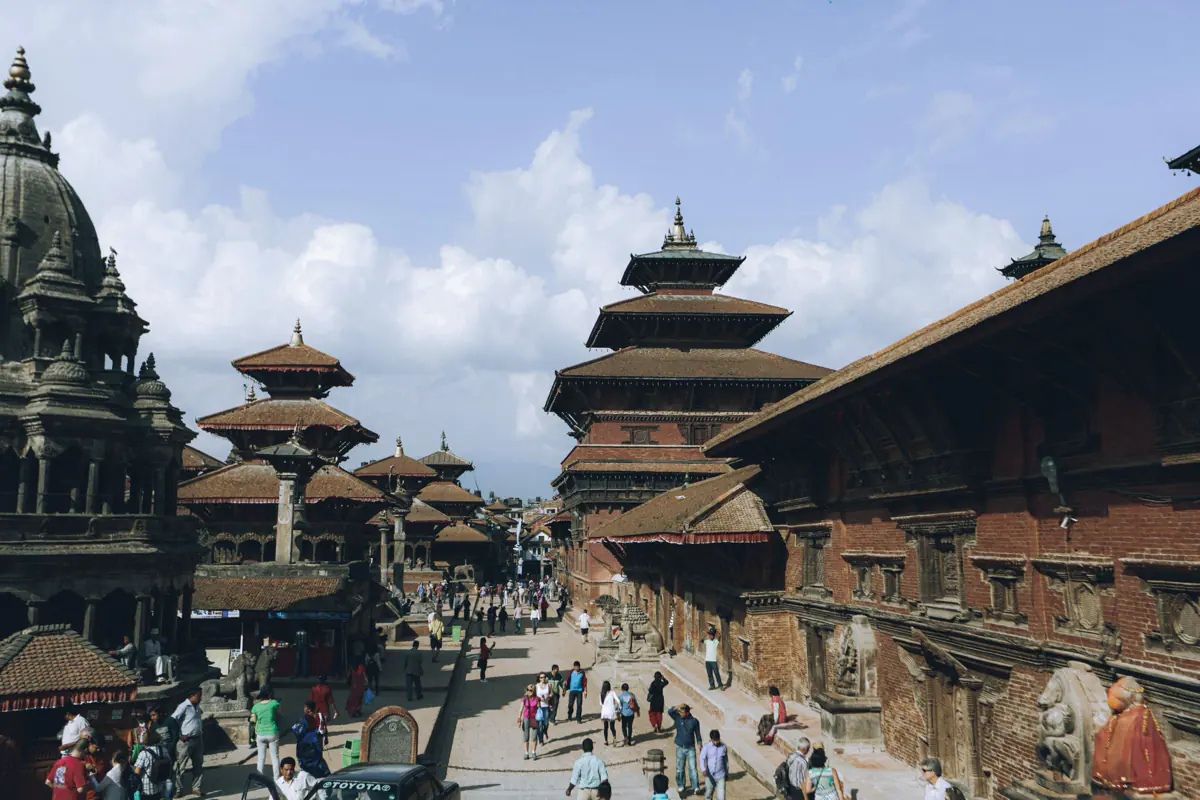 Tiered pagoda roofs and a carved stone temple around Patan Durbar Square, with visitors walking across the paved square